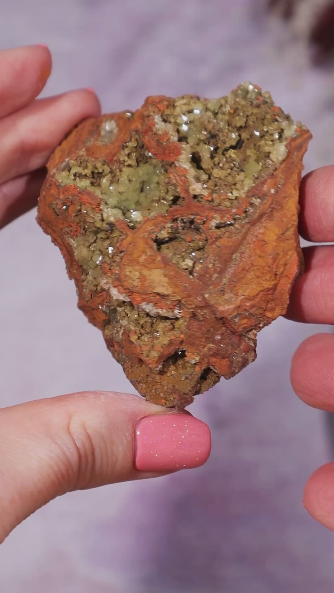 Adamite mineral held by a hand to show size between thumb and fingers. The specimen is turning it so you can see all sides of the rock and the deposits of crystals within the structure.